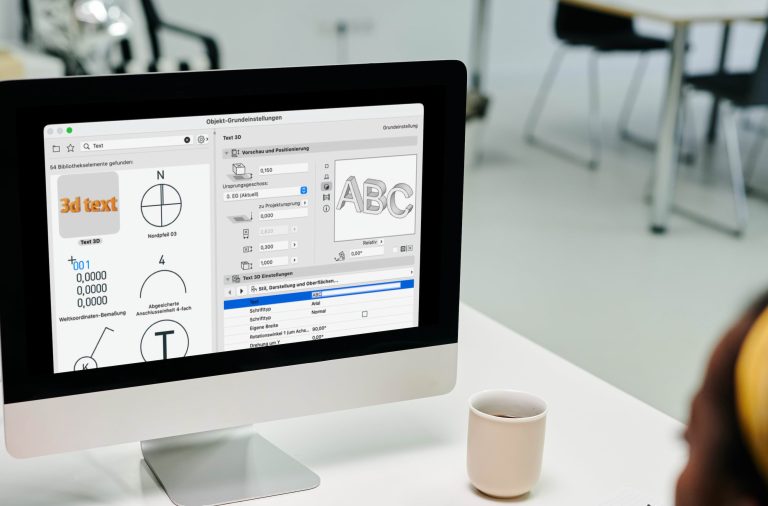 Ein Computermonitor zeigt eine Grafikdesign- oder CAD-Softwareoberfläche mit Archicad-Objekten und Textbearbeitungsfunktionen. Ein weißer Kaffeebecher steht in der Nähe auf dem Schreibtisch.