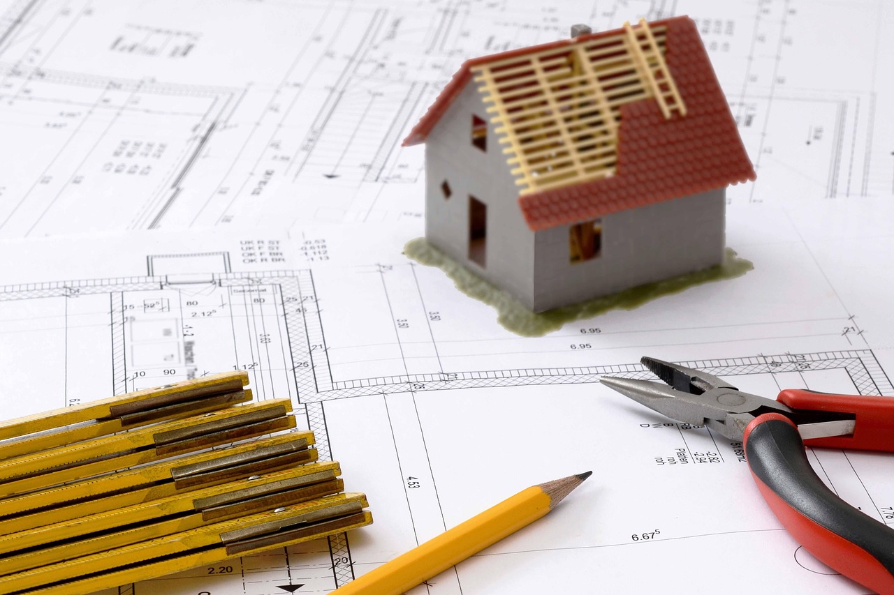 Auf den Bauplänen liegen Modellhaus, Zollstock, Bleistift und Zange – perfekte Tools für die digitale Planung oder ein entspanntes Bier+Brezeln-Brainstorming.