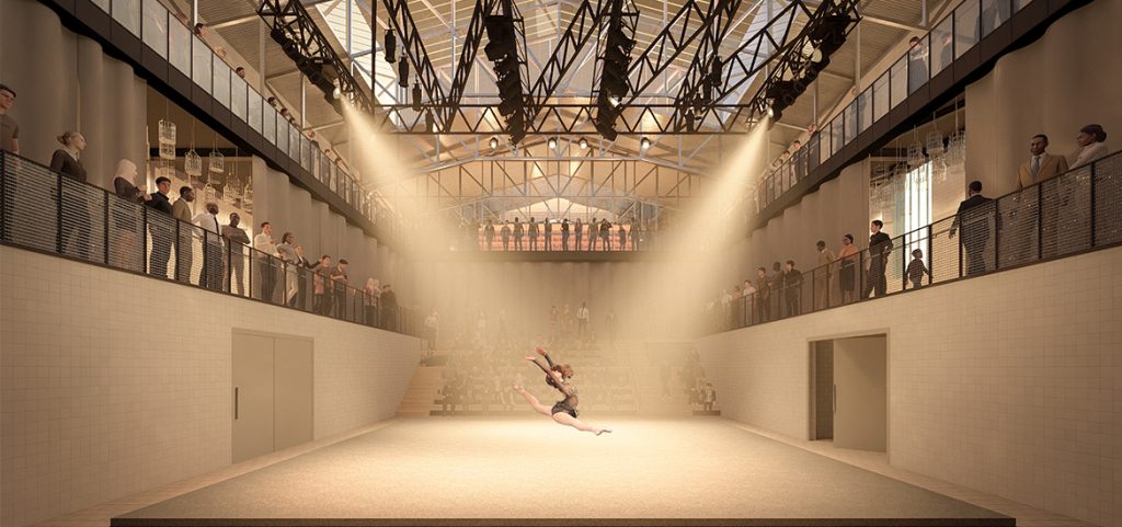 Eine Tänzerin springt in der Mitte einer gut beleuchteten Innenbühne, die von Zuschauern auf zwei Ebenen in dem von Maximilian Hofmann im industriellen Stil entworfenen Wandelwerk umgeben ist.