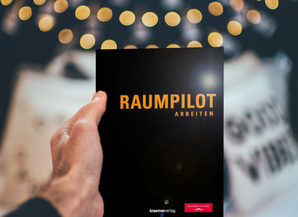 Eine Person hält ein Buch mit dem Titel „Raumplot“ hoch, das zum kostenlosen Download verfügbar ist.