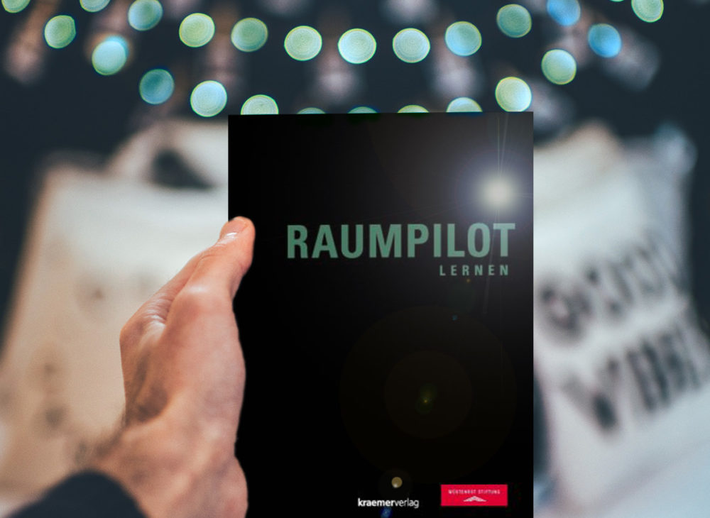 Eine Person hält ein Buch mit dem Wort „Raumpilot“ hoch, das als Lernressource kostenlos heruntergeladen werden kann.